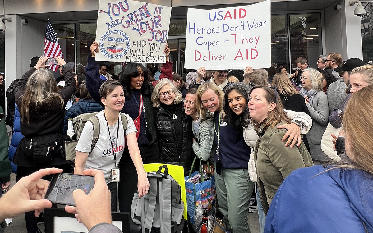 Crowd of USAID supporters posing outside with signs praising USAID employees and their aid work.